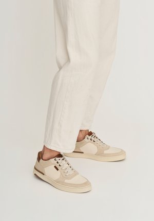 Persoon draagt beige en witte casual sneakers met een witte broek tegen een effen lichte achtergrond.