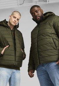 Giubbotti puffa verdi con design trapuntato, cappucci e cerniere. Due modelli che li indossano, uno con jeans chiari e l'altro con denim scuro.