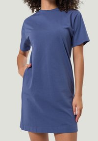 Femme portant une robe en coton bleue à manches courtes et longueur genou, avec sa main droite dans une poche latérale, sur un fond uni.