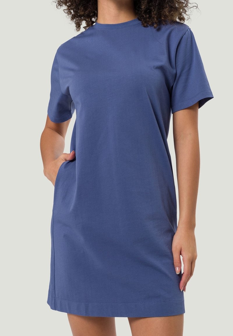 Femme portant une robe en coton bleue à manches courtes et longueur genou, avec sa main droite dans une poche latérale, sur un fond uni.