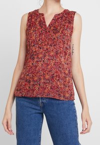 Ærmeløs bluse i et rødt blomstermønster med volangdetaljer ved halsudskæringen. Lavet af letvægtsstof. Sættes sammen med blå denimjeans.