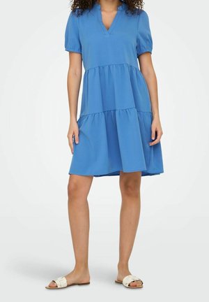 Femme portant une robe bleue ample à manches courtes arrivant au genou avec une jupe à volants et des sandales plates beiges sur un fond uni.