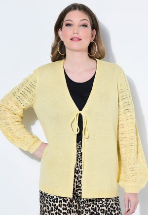 Kobieta ubrana w jasnożółty dziergany kardigan z rękawami balonowymi, czarny top, spodnie w cętki lamparta oraz duże kolczyki koła, stoi z ręką na biodrze.