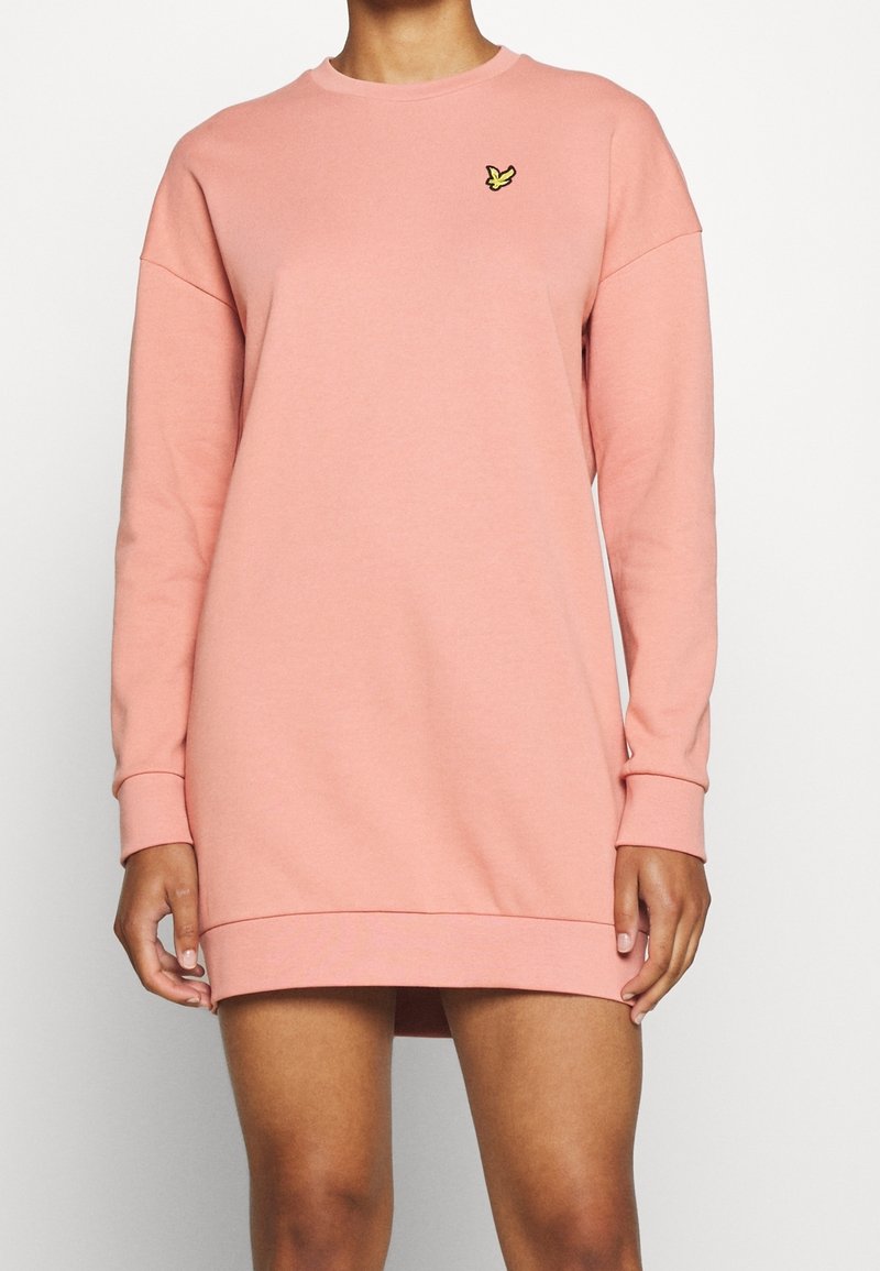 Robe sweat-shirt rose en tissu doux, avec manches longues et encolure ronde. Comprend un petit logo jaune sur la poitrine.