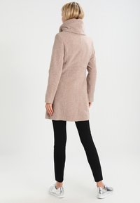 Cappotto beige in misto lana con collo alto a imbuto e vestibilità sagomata, abbinato a leggings neri e sneakers argentate, visto da dietro.