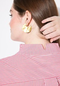 Orecchini con texture dorata e una camicia a righe rosse e bianche con colletto e dettagli di fessura posteriore, evidenziando gli elementi di tessuto e design.