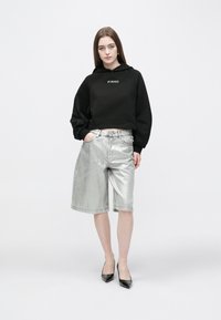 Cropped hoodie preto com o logótipo "PINKO", combinado com calções prateados brilhantes até ao joelho e saltos pontiagudos pretos, num fundo neutro.