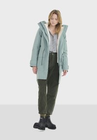 Parka bleu clair avec une capuche en polaire, fermeture éclair à l'avant et poignets ajustables, associée à un pantalon en velours côtelé vert foncé et des bottes noires.