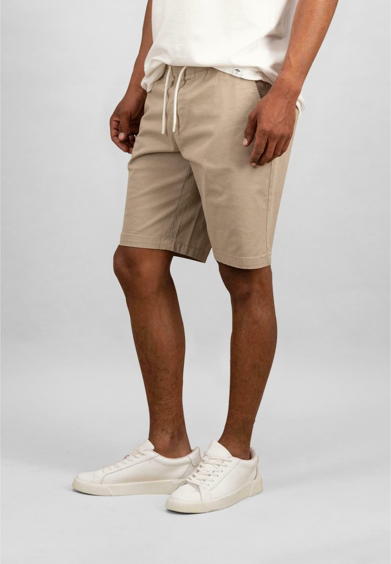 Beige shorts dengan pinggang elastis dan tali serut. Dipadukan dengan sepatu sneakers putih, memiliki desain rendah dan tali sepatu yang sederhana.