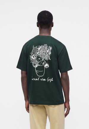 Person iført en mørkegrøn T-shirt med hvid linjetegning af solsikker i en vase og teksten "Vincent van Gogh" på bagsiden.