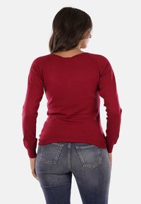 Maglione rosso lavorato a maglia con scollo a V, maniche lunghe e design aderente. La texture è liscia, abbinato a jeans di denim blu, per un look casual.