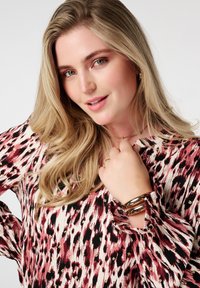 Blouse à manches longues dans un motif animalier rose, noir et crème, avec une coupe décontractée et des poignets froncés. Des accents de bijoux dorés sont visibles.