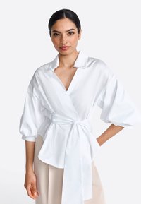 Weiße Wickelbluse mit V-Ausschnitt, gepufften Ärmeln und gebundenem Bund. Hergestellt aus glattem Stoff, zeichnet sich durch ein schlichtes Design und dezente Knöpfe aus.