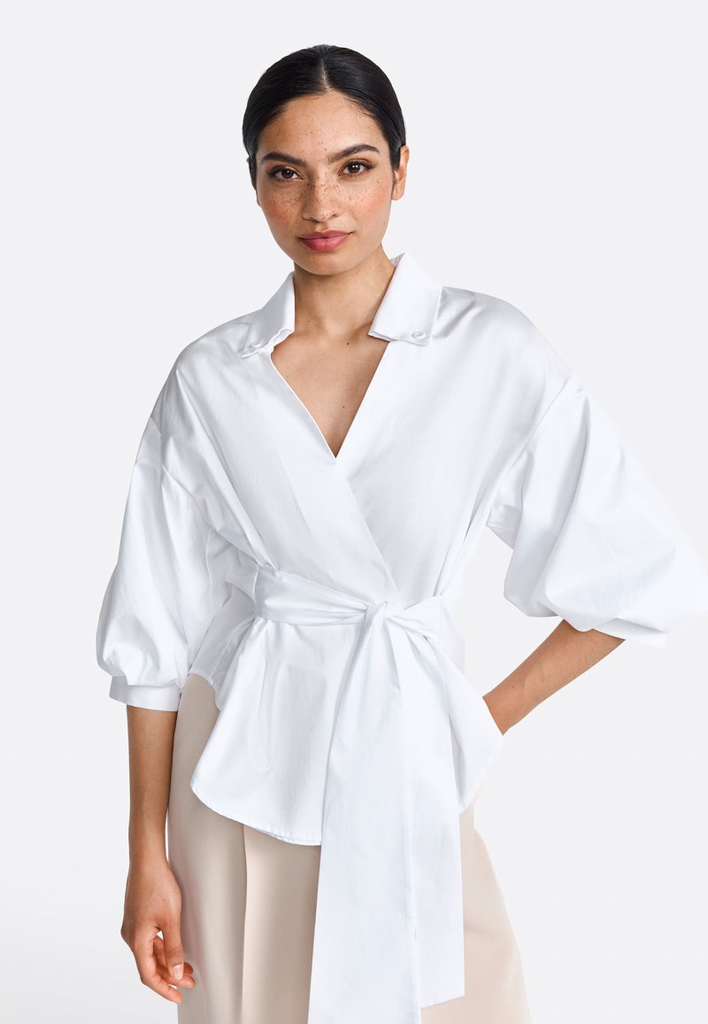Weiße Wickelbluse mit V-Ausschnitt, gepufften Ärmeln und gebundenem Bund. Hergestellt aus glattem Stoff, zeichnet sich durch ein schlichtes Design und dezente Knöpfe aus.