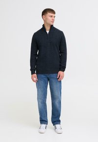 Marineblauwe half-zip sweater van zacht gebreid materiaal, gecombineerd met lichtblauwe jeans en witte sneakers met zwarte strepen. Het model staat zijwaarts gericht.