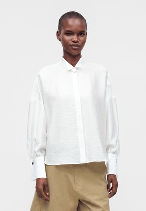 Persona con capelli corti che indossa una camicetta bianca a maniche lunghe e morbida e pantaloni beige, in piedi davanti a uno sfondo chiaro e uniforme.