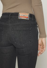 Jeans de mezclilla negra con un diseño ajustado, que cuentan con dos bolsillos traseros y una etiqueta de cuero color marrón con acentos rojos y azules en la cinturilla.