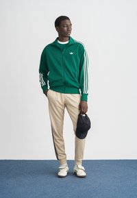 Zelená bunda na zips s bielymi pruhmi a logom, béžové tepláky s čiernymi bočnými pruhmi, biele tenisky a čierna čiapka držená v ruke.