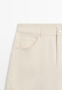 Pantaloni in denim beige a vita alta che mostrano la tasca anteriore sinistra, chiusura con bottone, passante per cintura e dettagli con rivetti in metallo su sfondo bianco.
