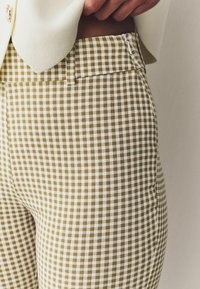 Pantalon à carreaux marron et blanc avec une texture lisse, doté d'une fermeture éclair avant et de détails de poche, présenté à côté d'un blazer de couleur crème.