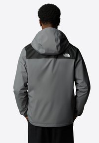 Grijze waterdichte jas met een zwart bovenpaneel en capuchon, voorzien van een logo op de linkerschouder en elastische manchetten. Soepele textuur.