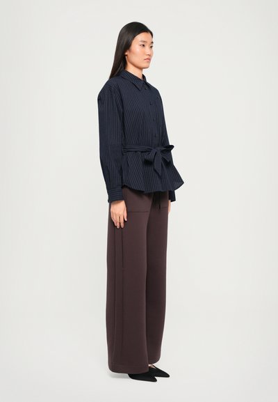 Navy striped button-up shirt with a belted waist and peplum design, paired with dark brown wide-leg trousers and pointed black shoes.