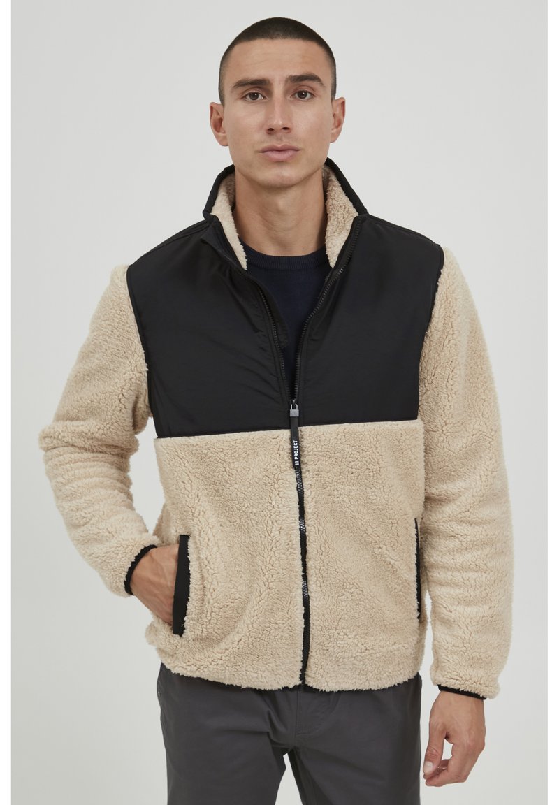 Tan and black fleece jacket with a zip front, sherpa texture on tan sections, and smooth fabric on black chest and sleeves. Features side pockets.