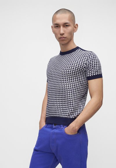 Homme à la tête rasée portant une chemise à manches courtes à motif bleu marine et blanc et un pantalon bleu vif, debout les mains dans les poches.