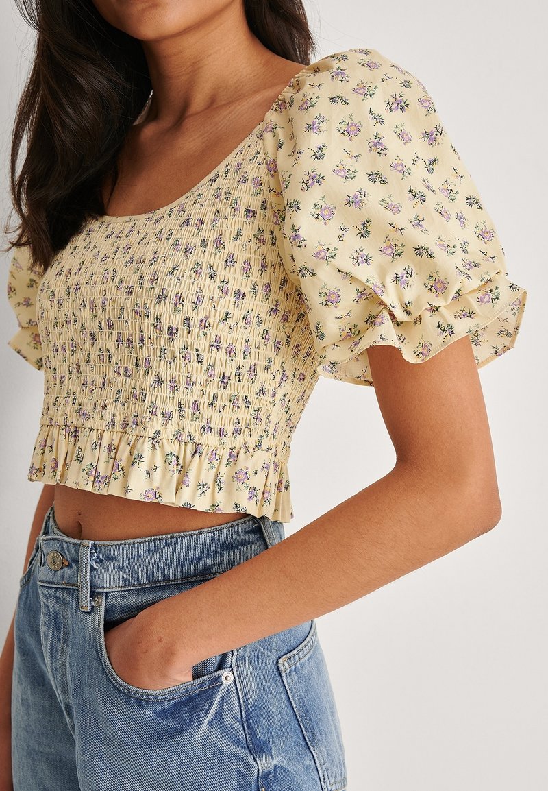 Crop top smock jaune avec de courtes manches bouffantes, à motif floral et ourlet volanté, porté avec un short en denim taille haute.