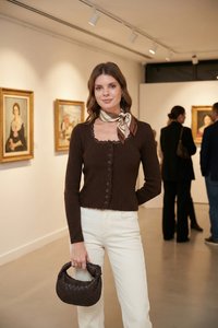 Young woman in white pants and brown buttoned sweater holds a woven bag in an art gallery with paintings and visitors in the background.