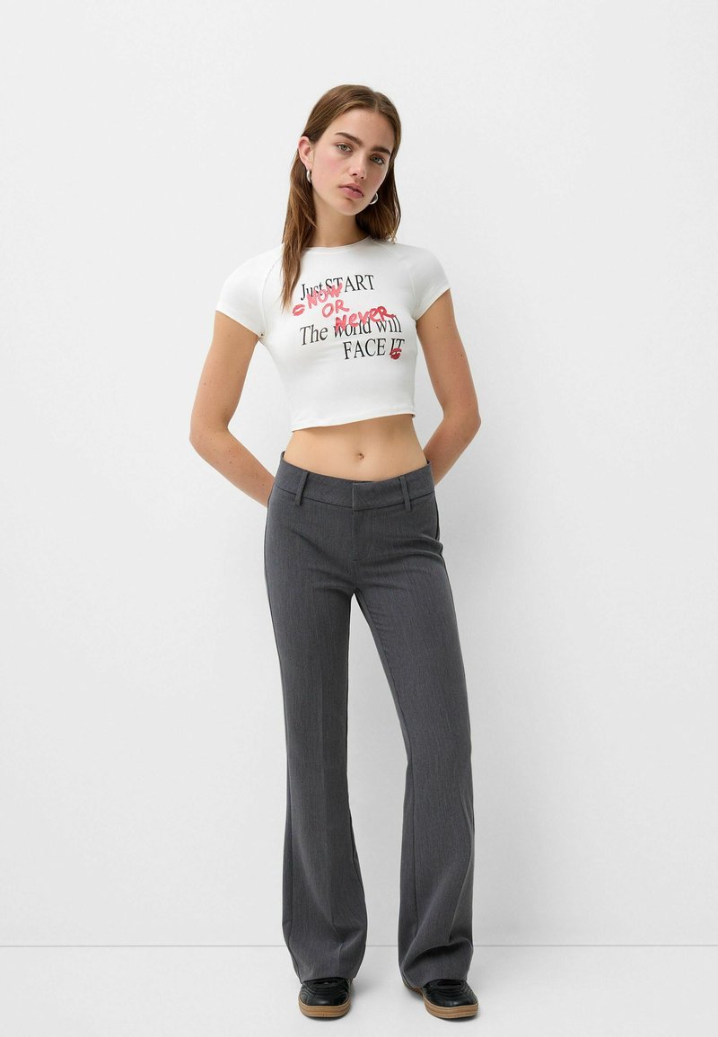 T-shirt bianca tagliata con stampa di testo nero e rosso; abbinata a pantaloni grigi svasati, con una texture liscia e una vestibilità su misura.