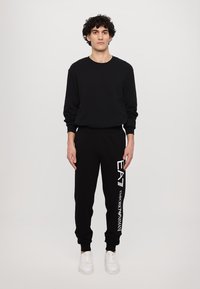 Čierna mikina s kruhovým výstrihom a predným vreckom, v kombinácii s čiernymi teplákmi s bielym logom "EA7" a textom "Emporio Armani". Biele tenisky.