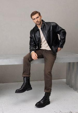 Czarna skórzana kurtka na kremowym swetrze, brązowe dżinsy i czarne kowbojki na sznurowadła. Tło jest szare, a scena zawiera metalową ławkę.