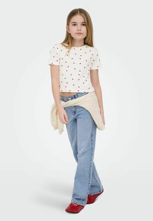 Ragazza giovane con lunghi capelli biondi che indossa un top corto bianco con puntini rossi, jeans azzurri, un maglione crema legato in vita e scarpe rosse.