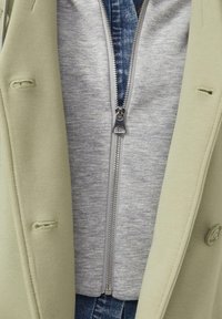 Nahaufnahme von geschichteter Kleidung mit einem beigen Mantel, einem hellgrauen Zip-up-Pullover und einem blauen Jeanshemd darunter.