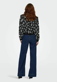 Femme aux longs cheveux ondulés, portant un pull à motifs noir et blanc, un jean bleu foncé à jambes larges et des chaussures à talons hauts noires, se tenant de dos.