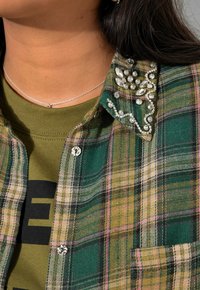 Groene en gele geruite flanellen shirt met kristallen versieringen op de kraag, met metalen knopen, en gelaagd over een olijfgroen shirt.