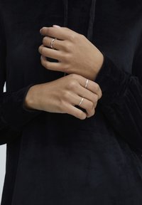 Silberne Ringe, die an Händen getragen werden, die ein schwarzes, samtiges Oberteil tragen. Die Ringe umfassen ein verdrehtes Design und ein schlichtes Band, die ein elegantes Finish zeigen.