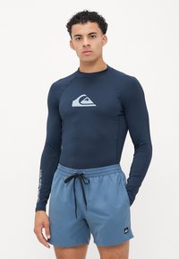 Tengeri kék hosszú ujjú rash guard világoskék logóval, világoskék fürdőnadrággal párosítva, amelynek húzózsinórja van a derekán és logófolt található az oldalán.