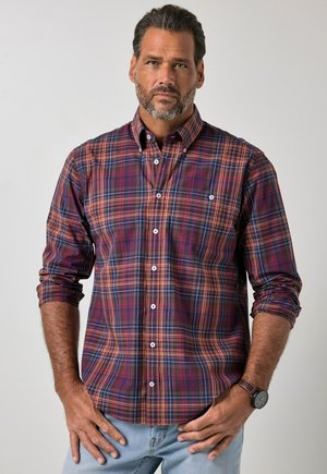 Man with short beard wearing a long-sleeve red, orange, and blue plaid shirt and light blue jeans, standing with hands near waist.