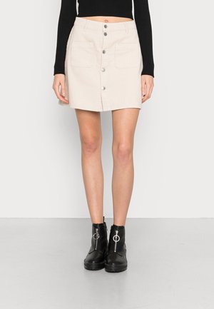 Beige corduroy rok met een rechte snit, gecentreerde knoopsluiting en twee zakken aan de voorkant. Draagt met zwarte enkellaarsjes met ritsen.
