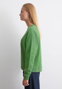 Grön sweatshirt med rund hals och långa ärmar, tillverkad av slät tyg. Har ribbade ärmslut och fåll, bärs tillsammans med mörkblå jeans.