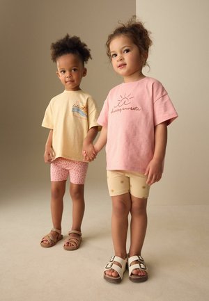 Doi copii se țin de mână, purtând tricouri și pantaloni scurți în nuanțe pastel cu sandale, stând pe un fundal bej simplu.