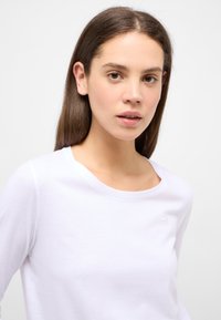 Weißes Langarm-T-Shirt mit rundem Halsausschnitt und einem kleinen, dezenten Logo-Detail auf der Brust. Weiches Baumwollmaterial mit glatter Textur.