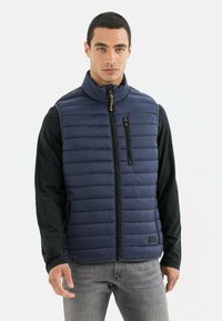 Marineblauwe gewatteerde puffer vest met ritssluiting aan de voorkant, hoge kraag, ritszakken en een logo-accent. Draagt over een zwart langemouwen shirt en grijze jeans.