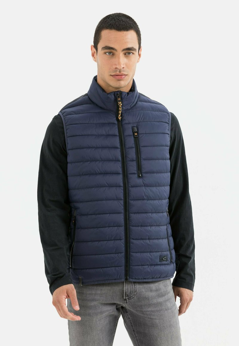 Marineblauwe gewatteerde puffer vest met ritssluiting aan de voorkant, hoge kraag, ritszakken en een logo-accent. Draagt over een zwart langemouwen shirt en grijze jeans.