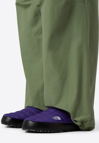 Lila isolerade tofflor med ett quiltat mönster och svarta gummisulor, kombinerade med olivgröna byxor, vilket skapar en lager på lager-look.