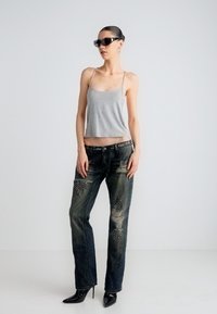 Hall hall lõigatud tank-top, kulunud tumedad teksad naelutatud aktsentidega ja mustad stiletto-saapad. Modell kannab tumedaid päikeseprille ja seisab valge tausta ees.