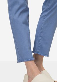 Světle modré džíny s třepeným lemem, slim fit a viditelným šitím. Nosené s krémovými šlapkami na neutrálním pozadí.