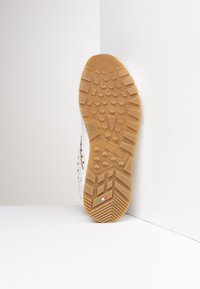 Gummisohle mit einem braunen strukturierten Grip-Muster, kombiniert mit einem weißen Obermaterial, das über perforierte Details und eine auf der Seite eingestanzte Markenkennzeichnung verfügt.